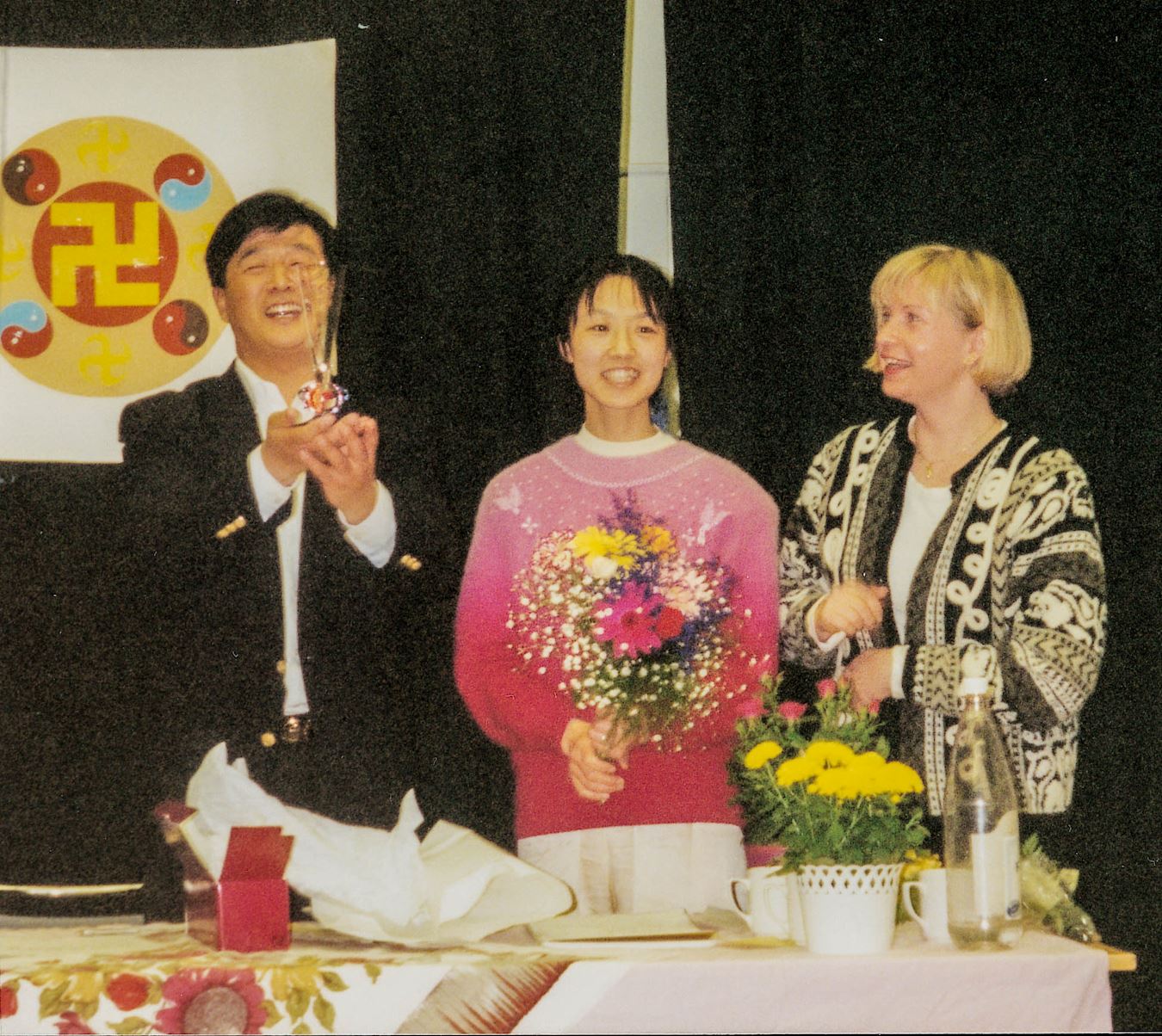 '图2:李洪志师父(左)高兴的向大家展示学员赠送的精美的小纪念品,王医生(中)站在师父旁边,帮师父拿着学员首先向师父敬献的鲜花。'